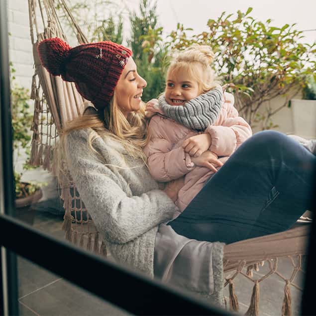 Junge blonde Frau mit roter Wintermütze und Tochter liegen in einer Hängematte auf dem Balkon und lächeln.