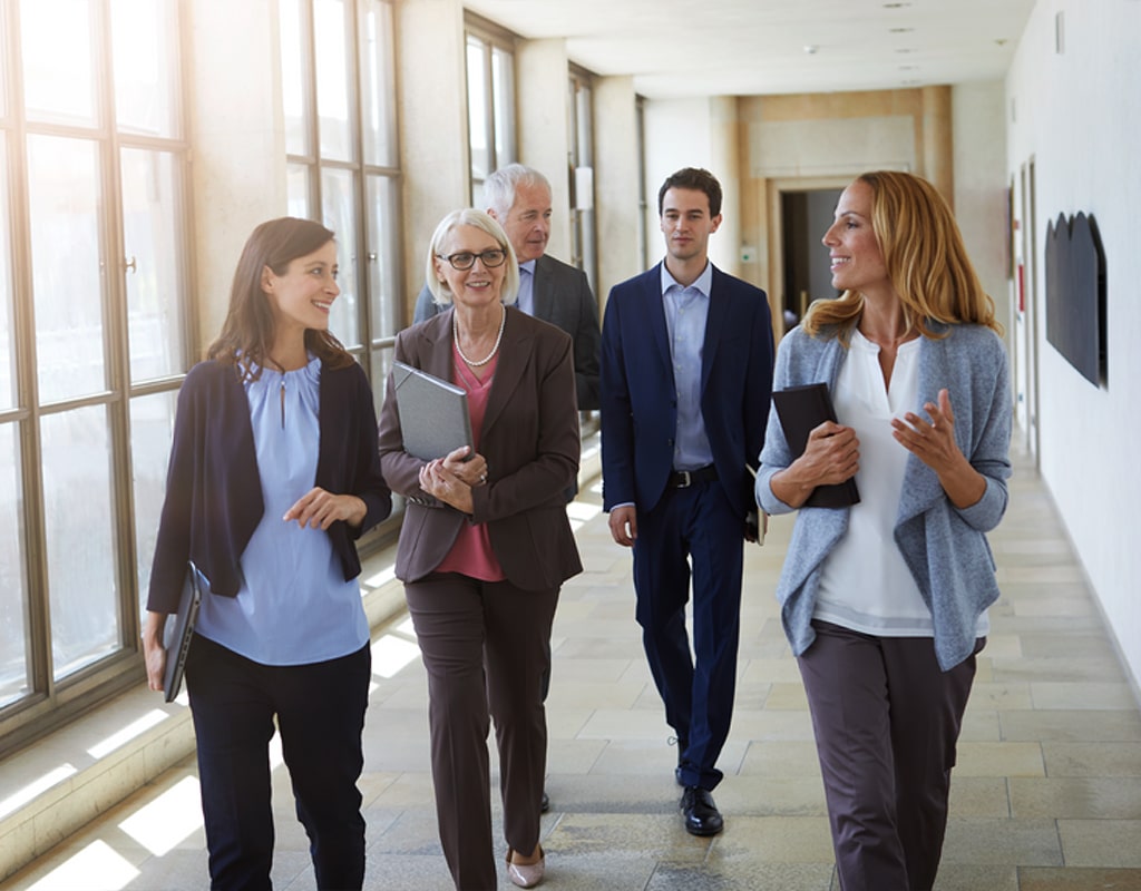 Gruppe von fünf Immobilienmaklern in Businesskleidung geht durch einen Flur. Die Frauen interagieren miteinander und lächeln, während der ältere Mann den jüngeren anschaut.