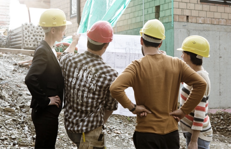 Vier Bauarbeiter*innen studieren einen Bauplan, während im Hintergrund ein Haus entsteht. Fleißige Arbeit am zukünftigen Wohnort.
